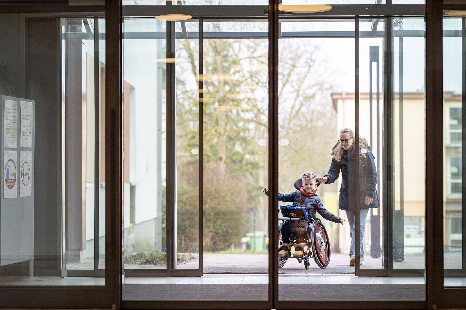 Kind im Rollstuhl betritt mit seiner Mutter das SPZ