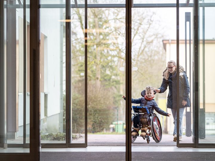Kind im Rollstuhl betritt mit seiner Mutter das SPZ