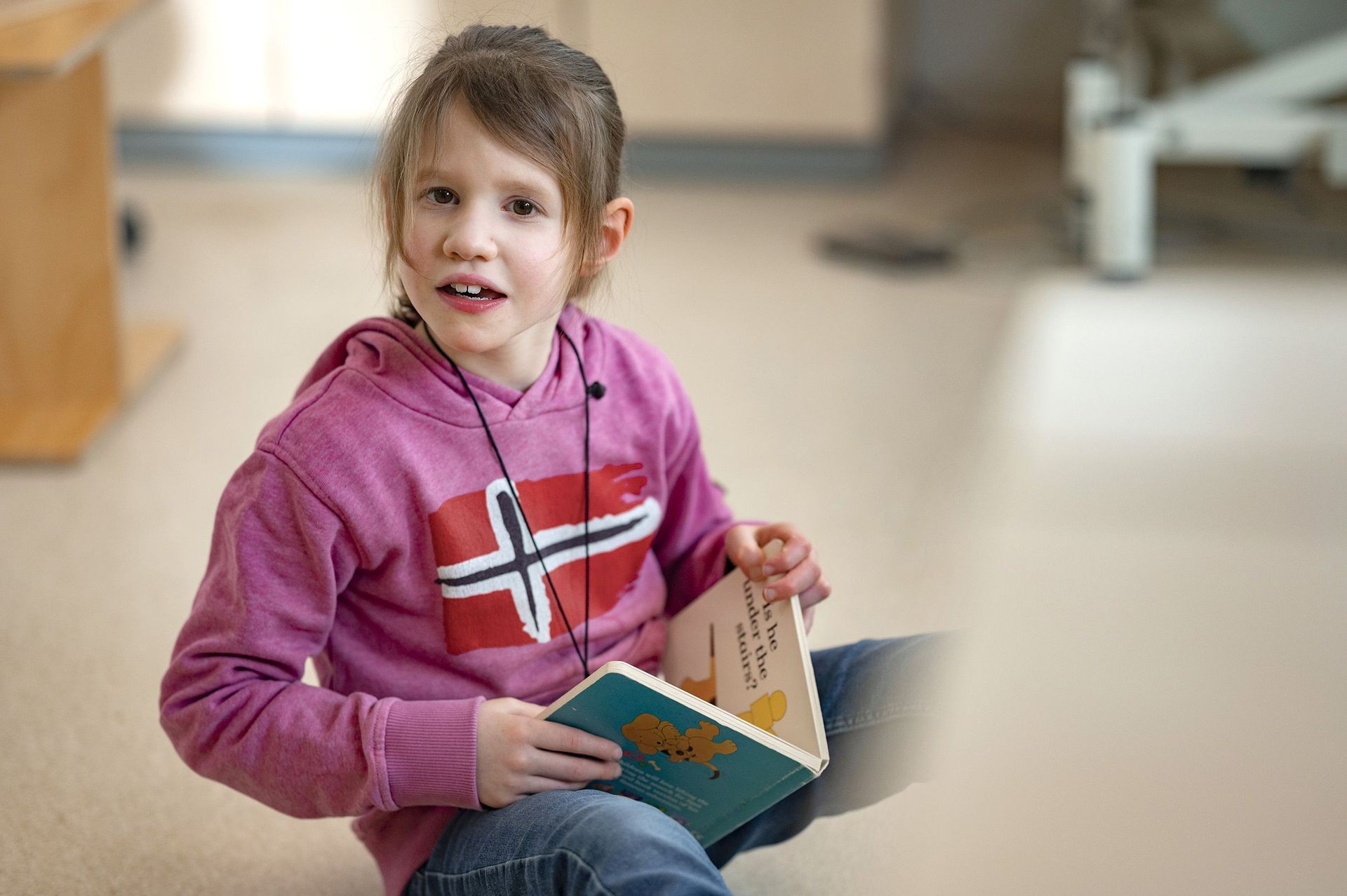 Kind sitzt auf dem Fußboden, liest in einem Buch und schaut in die Kamera 