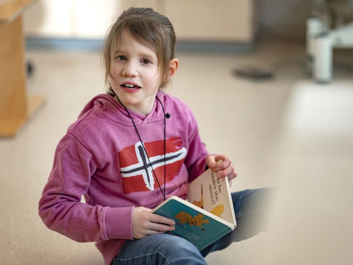 Kind sitzt auf dem Fußboden, liest in einem Buch und schaut in die Kamera 